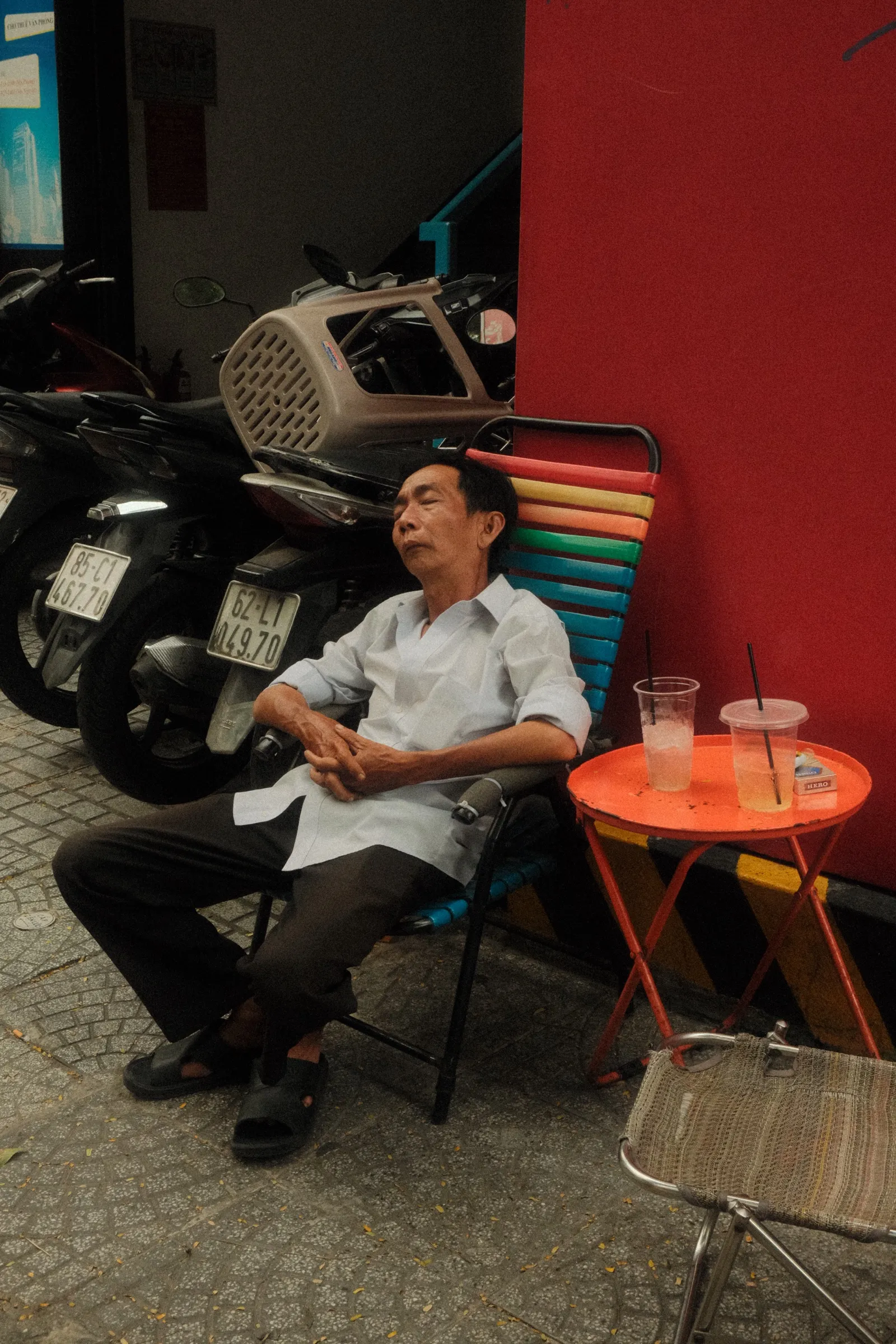 A man asleep upright at a cafe table in Hanoi, two empty plastic cups in front of him.