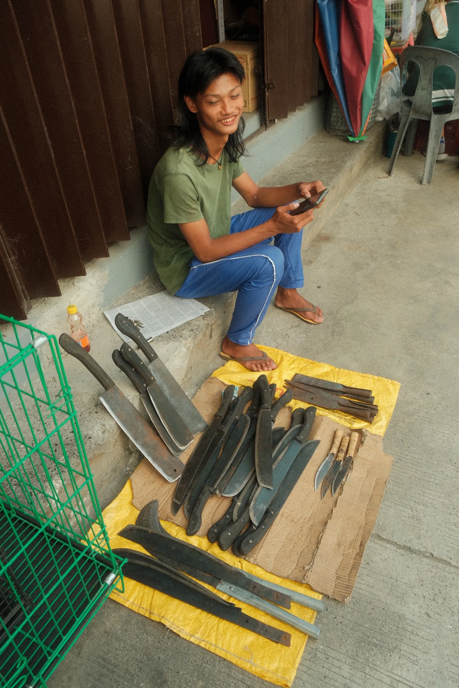 Machete salesman sitting on the curb in Bontoc behind a yellow tarp piled with bolos.